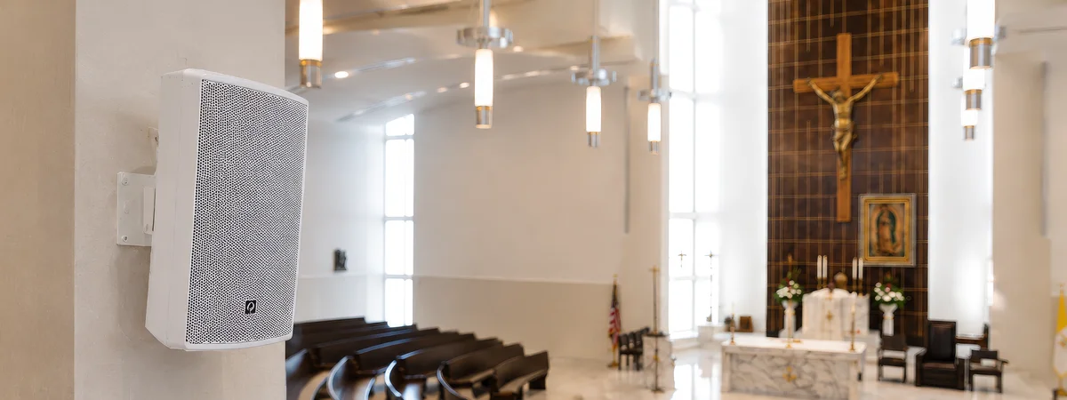 White column-mounted church speaker inside a modern sanctuary with curved pews, pendant lighting, altar, crucifix, and worship audio system integration